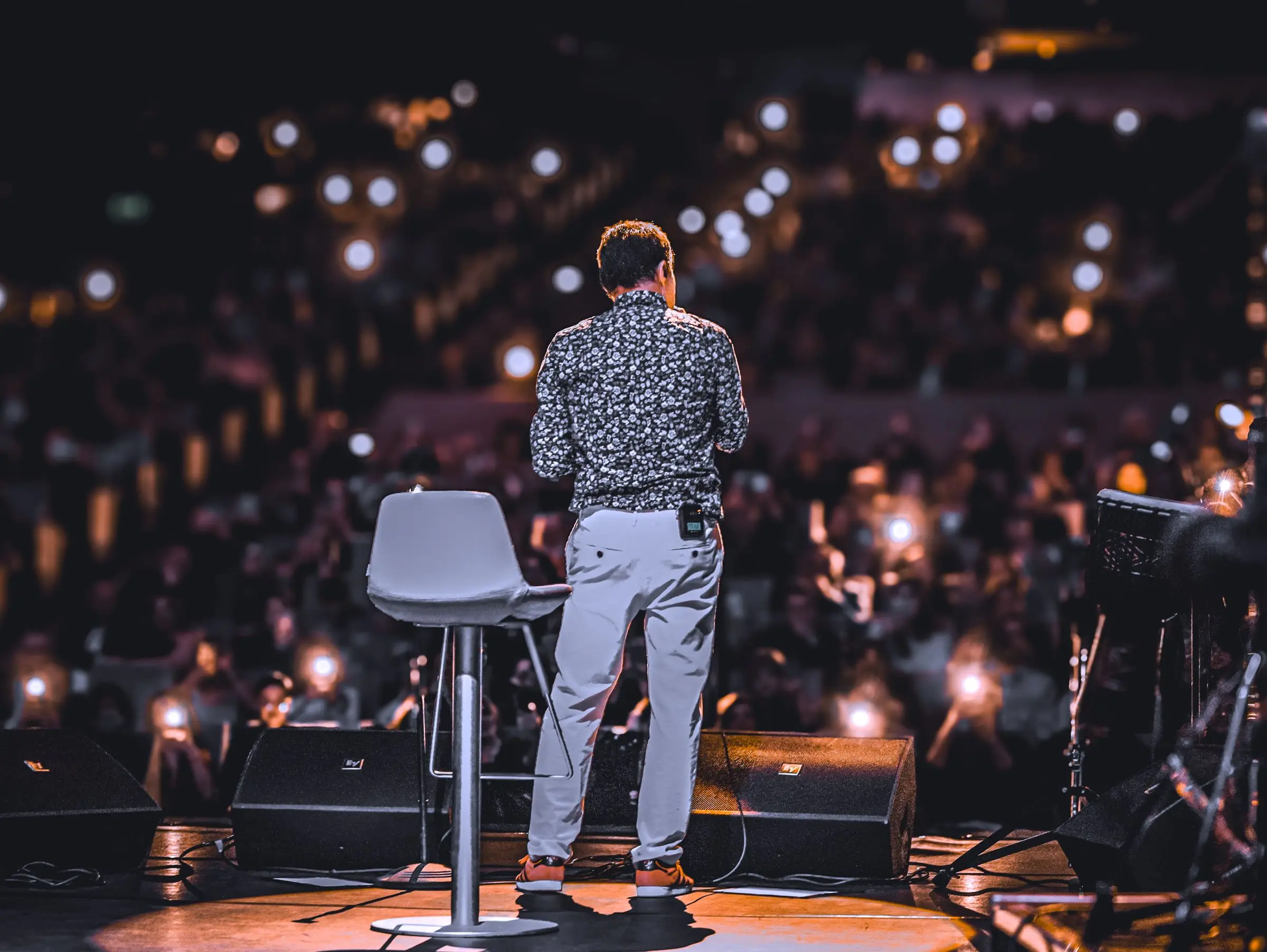 A speaker standing at the edge of a stage, facing a full auditorium lit by phone screens